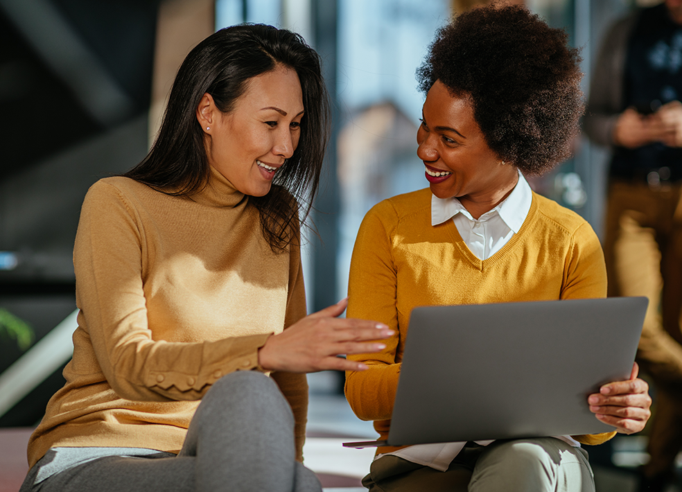 Two women sitting on a bench talking to each other. The woman on the right has a laptop in her lap.