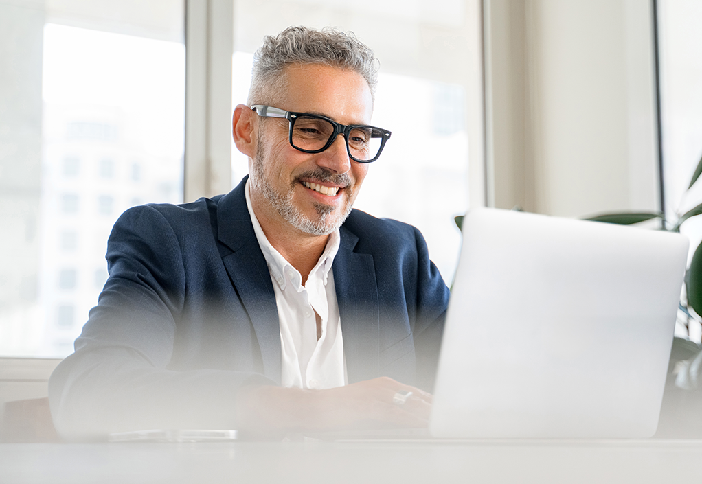 A professional man in a suit sitting at a desk, typing on a laptop and smiling.