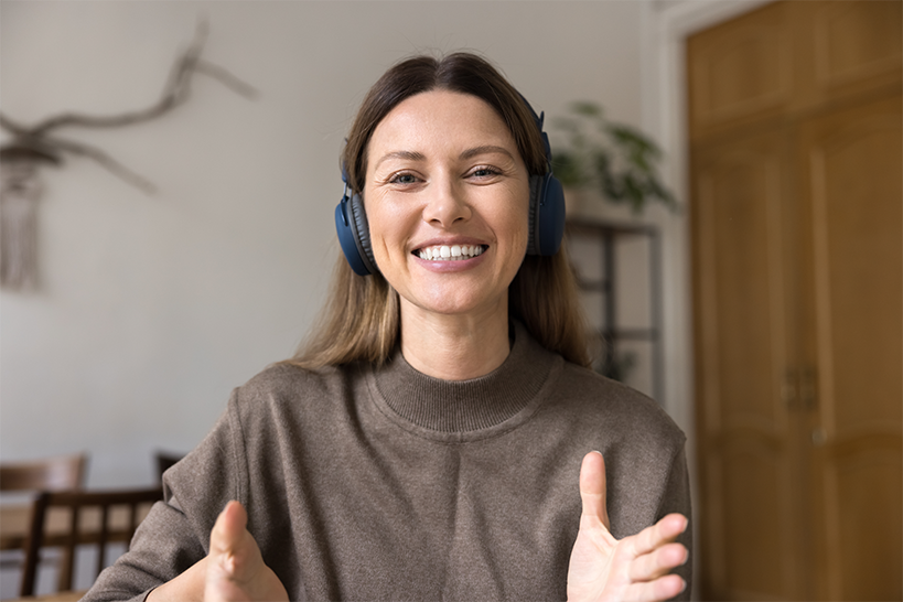 A woman in a sweater and over the head headphones looking at the camera smiling with her hands out as she is talking.