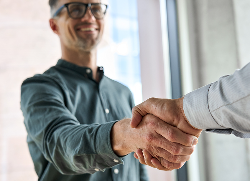 The focus is of two hands coming together for a handshake, with a smiling professional looking man out of focus and the other hand out of frame.