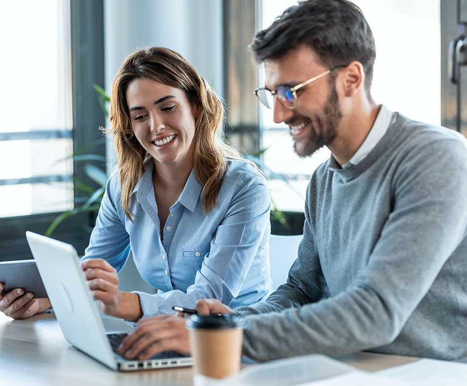 Two young professionals sitting at a desk. A smiling man is in front of a laptop, adjusting the screen and a woman is next to him holding a tablet and smiling.