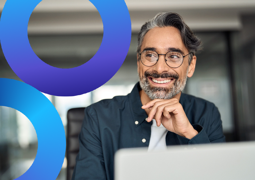 A man sitting at a desk in front of a laptop, smiling and looking off camera with his hand under his chin.