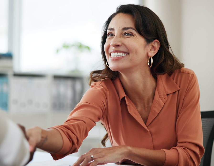 A woman, standing with arms crossed in business attire, smiles and looks through a glass window.