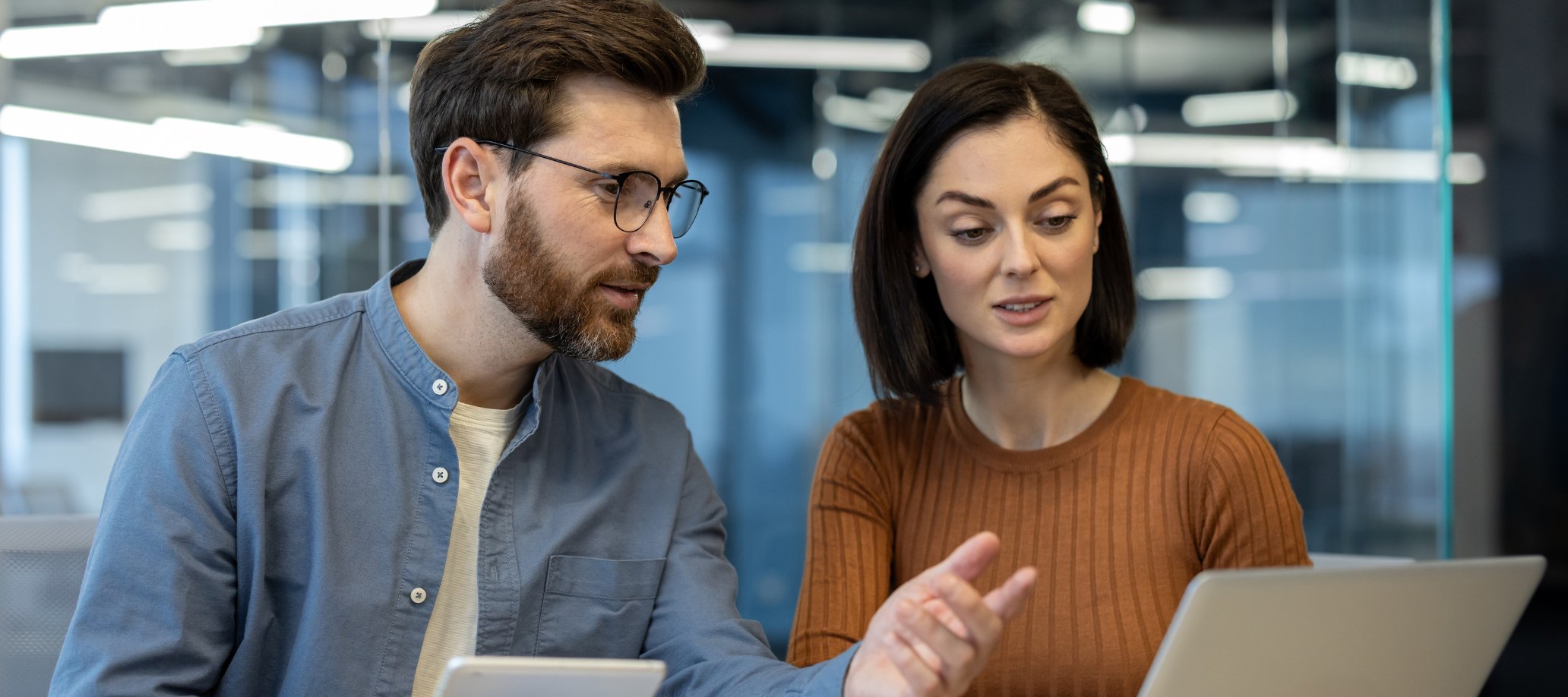 A man and a woman sitting at a desk, collaborating together. The man is holding a tablet and pointing at the laptop in front of the woman.