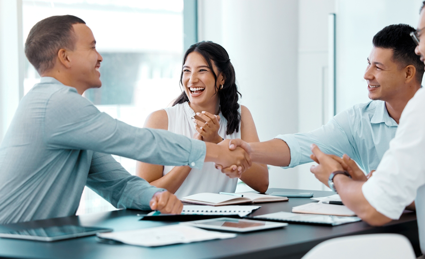 Group of four professionals sitting around a table, two men across from each other are shaking hands and a woman behind them is clapping her hands together.