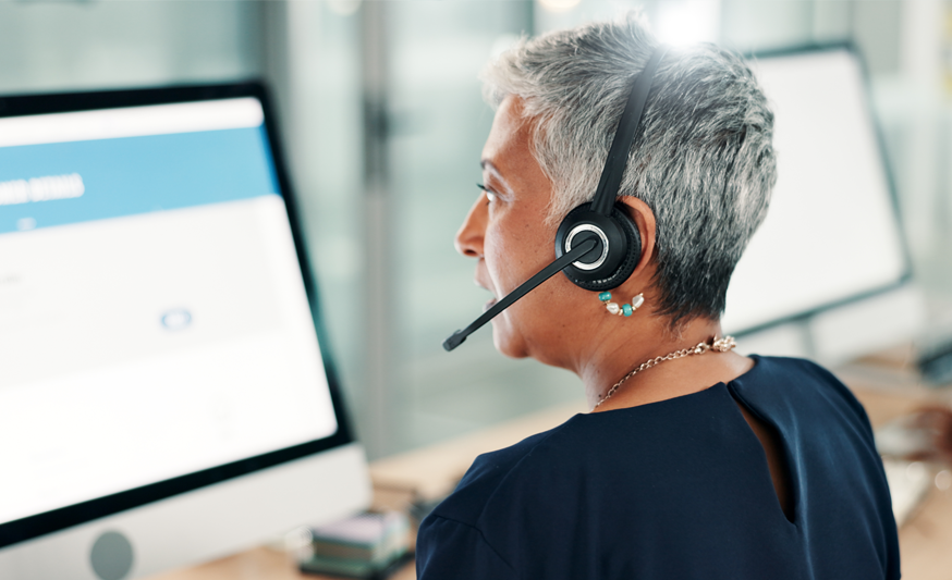 A woman is sitting in front of a computer screen, with her back to us and a headset on with a microphone.p