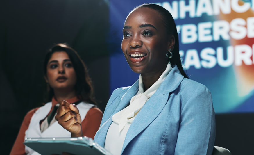 Two women sitting on a stage with a screen behind them. The one woman closest to the camera is the focus, she's smiling and holding a tablet.