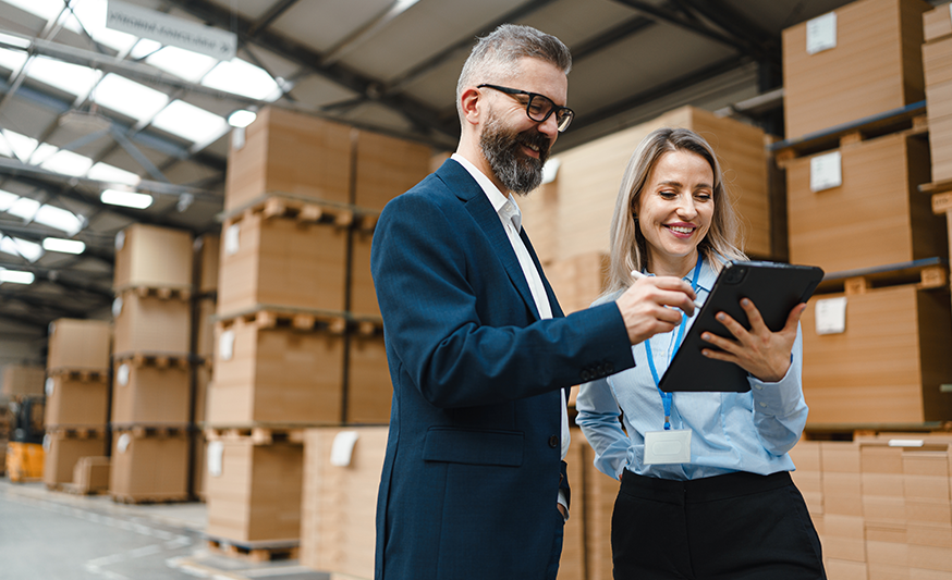 A man and a woman standing in a warehouse full of boxes, the woman is holding a tablet and the man is signing it.