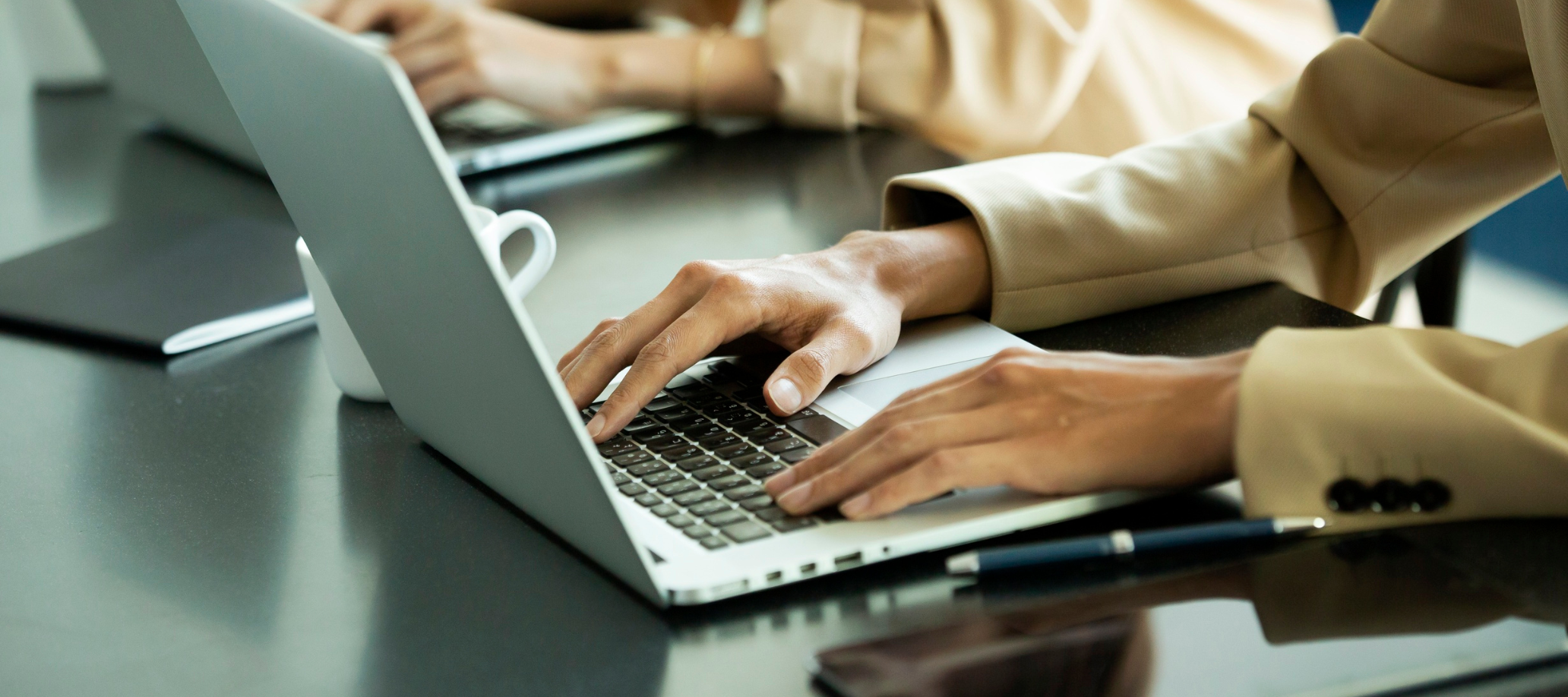 Two people sitting a desk with laptops open, the photo is focusing on their hands as they type on their keyboards.