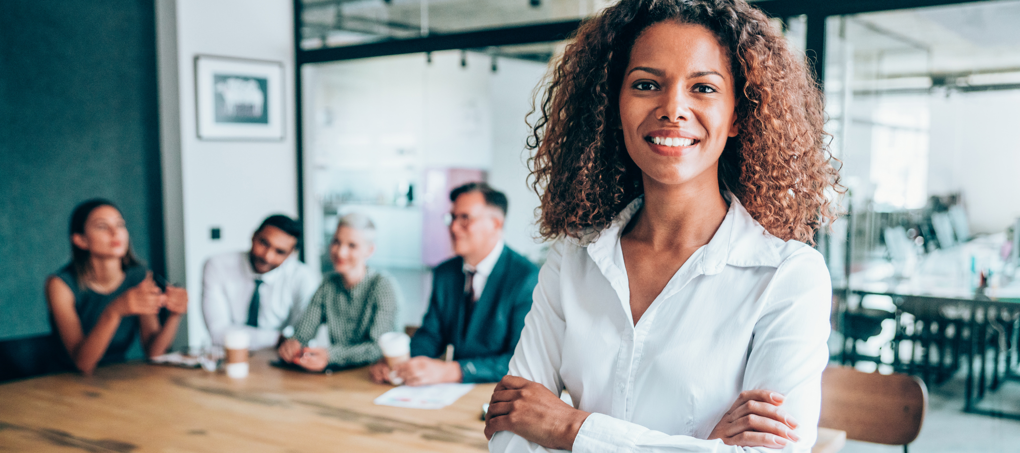 A woman leader is standing in front of a table with other professionals behind her. She is smiling at the camera with her arms crossed.