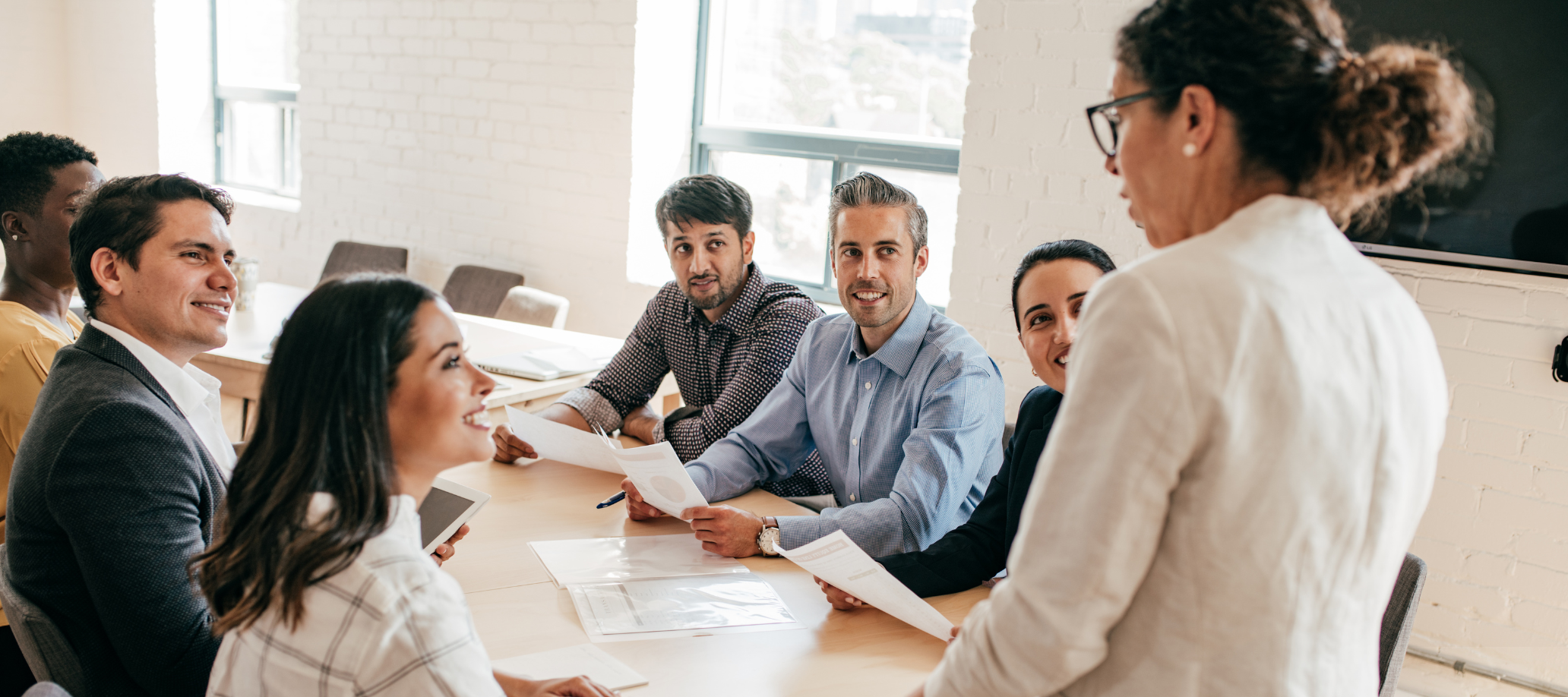 A group of professionals sitting at a table, looking at one woman who is standing at the head of the table with her back facing the camera.