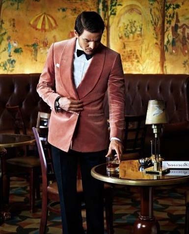 Un serveur élégant en veste saumon et nœud papillon noir pose une boisson sur une table dans un décor de restaurant vintage.