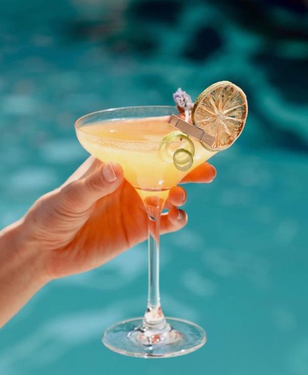 Main tenant un cocktail jaune dans un verre à pied avec une tranche de citron séché et une décoration, devant une piscine bleue floue en arrière-plan.
