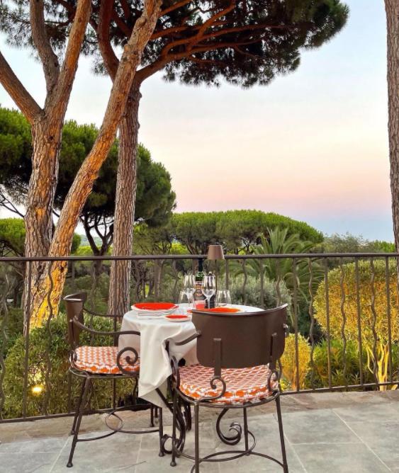 Une table de terrasse dressée pour deux personnes avec des assiettes rouges, sous des grands arbres au coucher du soleil.