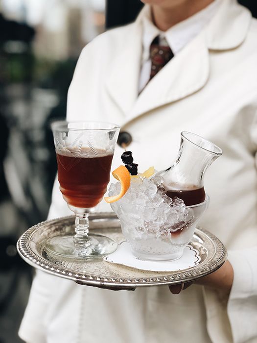 Serveur en veste blanche portant un plateau avec un verre de boisson brune et une petite carafe posée dans un bol de glace avec une garniture d'orange.