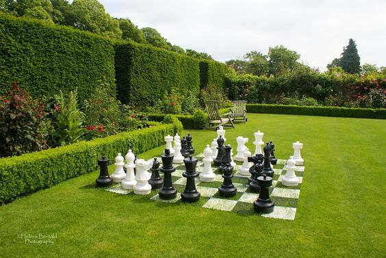 Échiquier géant avec pièces noires et blanches installé sur une pelouse verte dans un jardin avec haies taillées et deux chaises en bois.