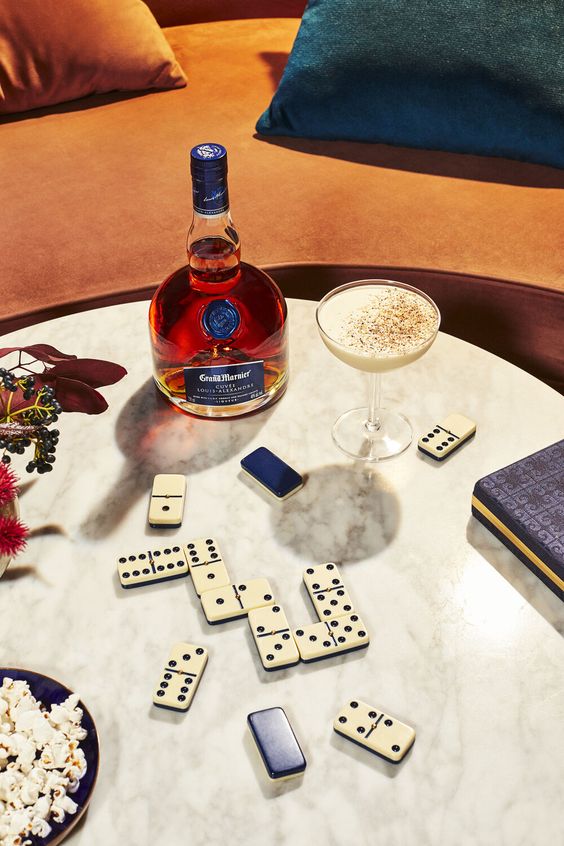 Bouteille de Grand Marnier, verre de cocktail, dominos et bol de pop-corn sur une table en marbre à côté d'un canapé avec coussins colorés.