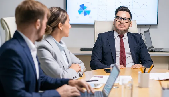Three business professionals in a meeting room, one man speaking while others listen attentively with charts displayed on a screen behind.