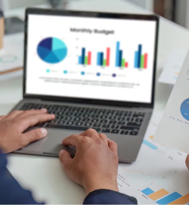 Person working on a laptop displaying colorful pie and bar charts labeled Monthly Budget, with printed charts on the desk.