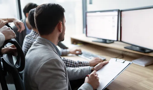 Business professionals collaborating at a desk with dual computer monitors, one person holding a clipboard.