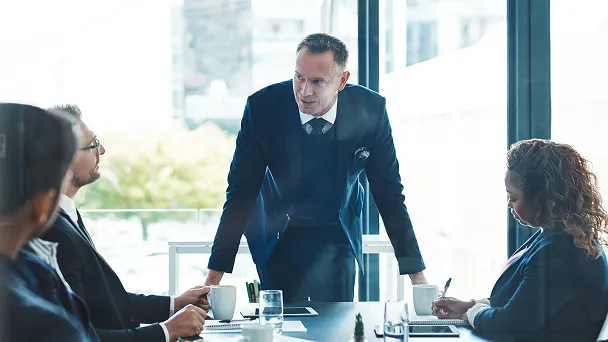 Businessman standing and speaking to colleagues seated around a conference table in a modern office.