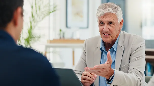 Older man in a beige blazer talking and gesturing with an unseen person across a table in a bright office.