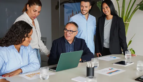 Five diverse colleagues collaborate around a laptop in a modern office meeting.