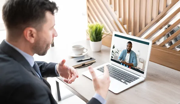 Man in a suit gesturing during a video call on a laptop showing another man on screen.