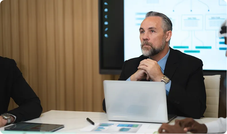 Bearded man in a suit sitting at a conference table with a laptop, attentively listening during a meeting.