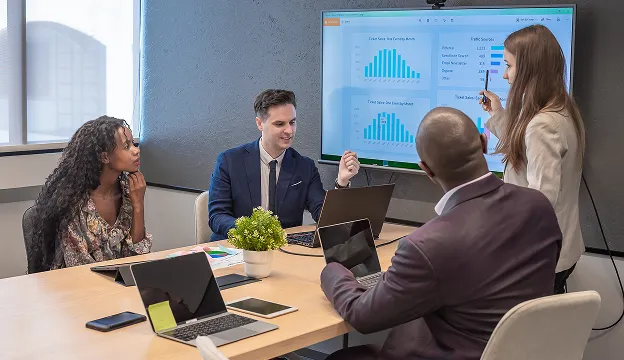 Four diverse colleagues in a meeting room discussing data charts displayed on a large screen.
