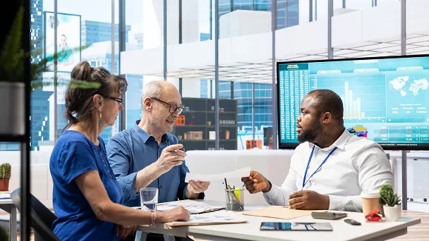 Three colleagues discussing documents in a modern office with a large screen displaying data charts.