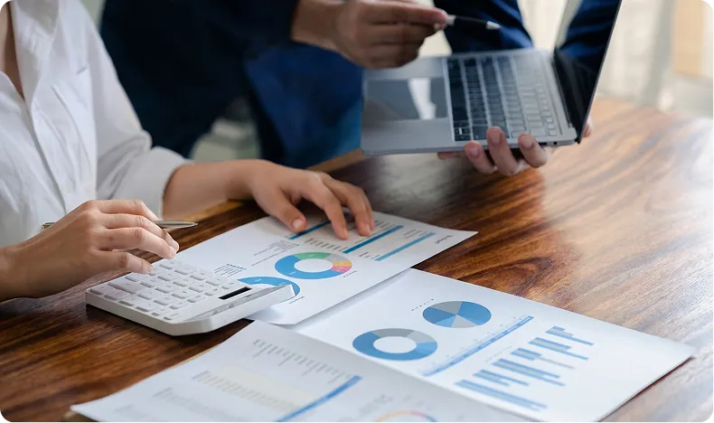 Two people reviewing financial charts and graphs with a calculator and a laptop on a wooden table.