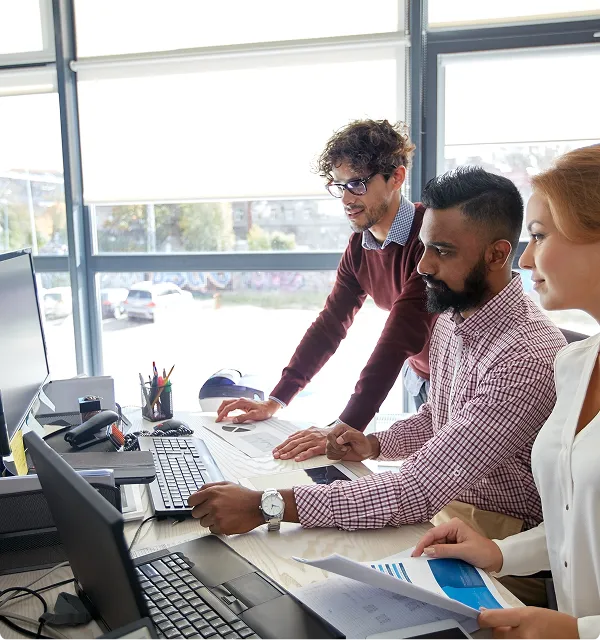 Three professionals collaborating and reviewing documents together at a desk with computers in a bright office.