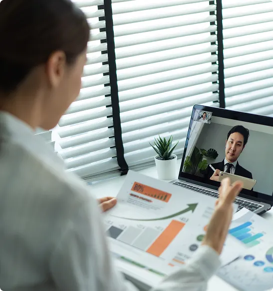 Person holding a paper with charts in front of a laptop showing a man in a suit during a video call.