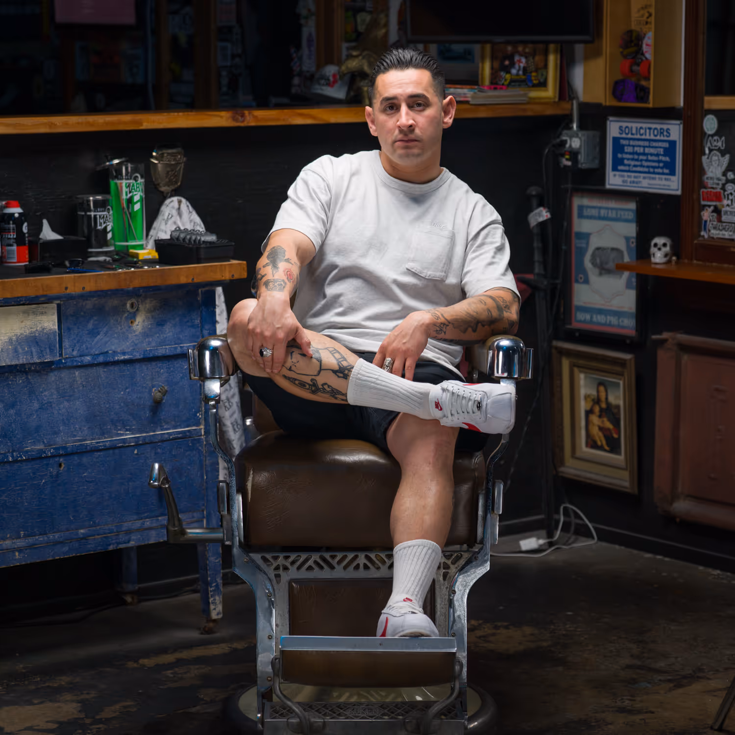 Man with tattoos sitting cross-legged on a vintage barber chair in a barbershop.