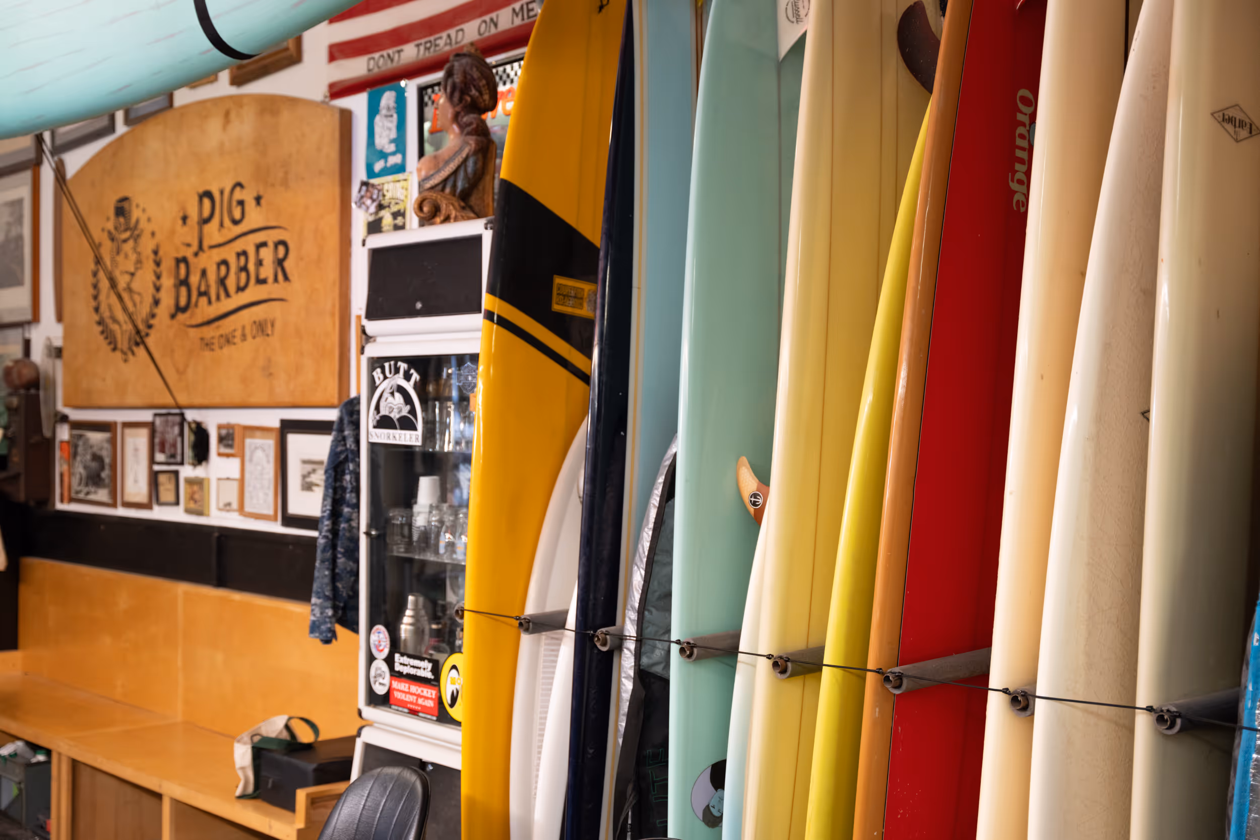 Colorful surfboards lined up vertically inside a barbershop with a wooden sign reading 'PIG BARBER' on the wall.