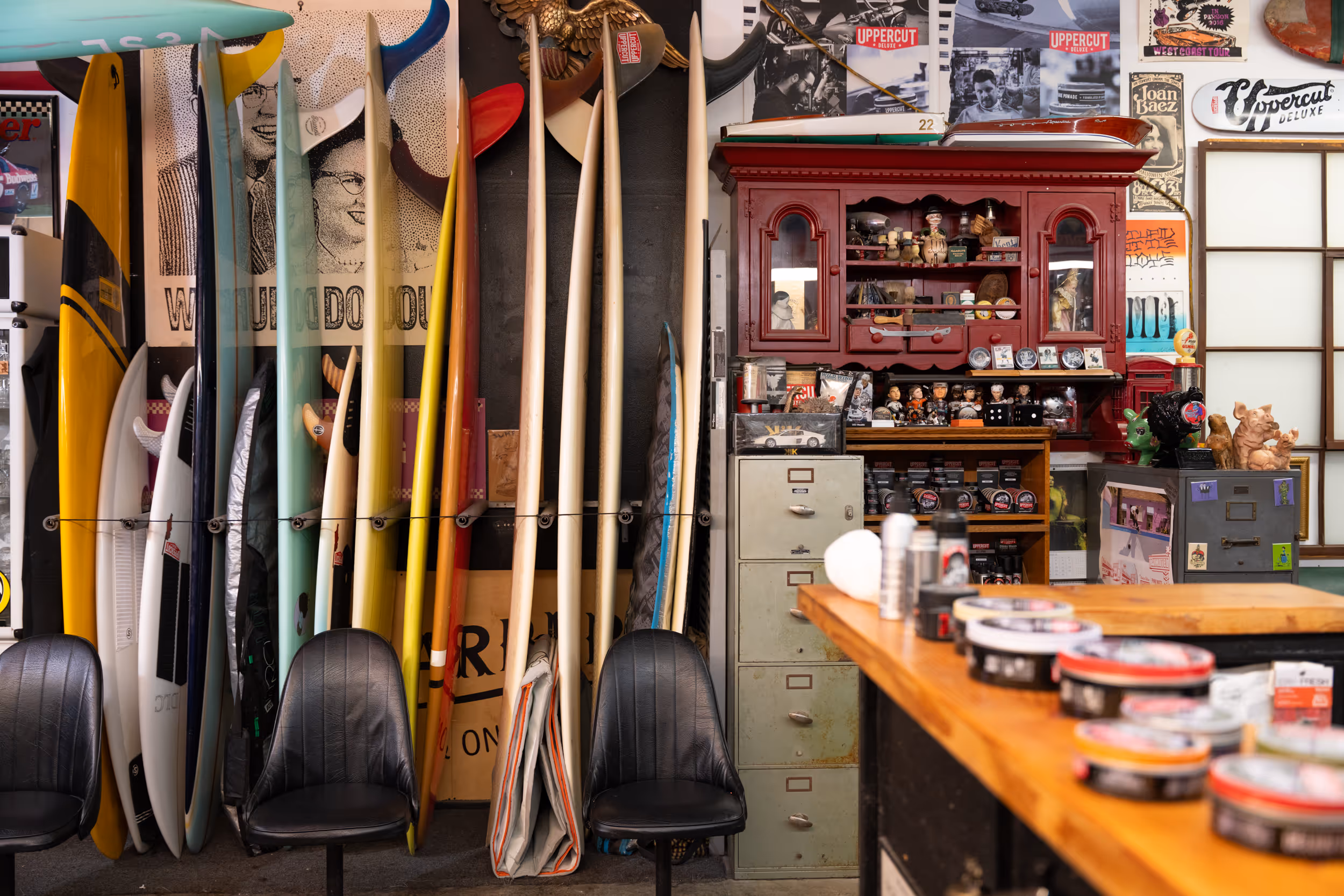 A row of colorful surfboards standing upright between two black chairs in a vintage-style barbershop with a red wooden cabinet, filing drawers, and various decorations on the walls.