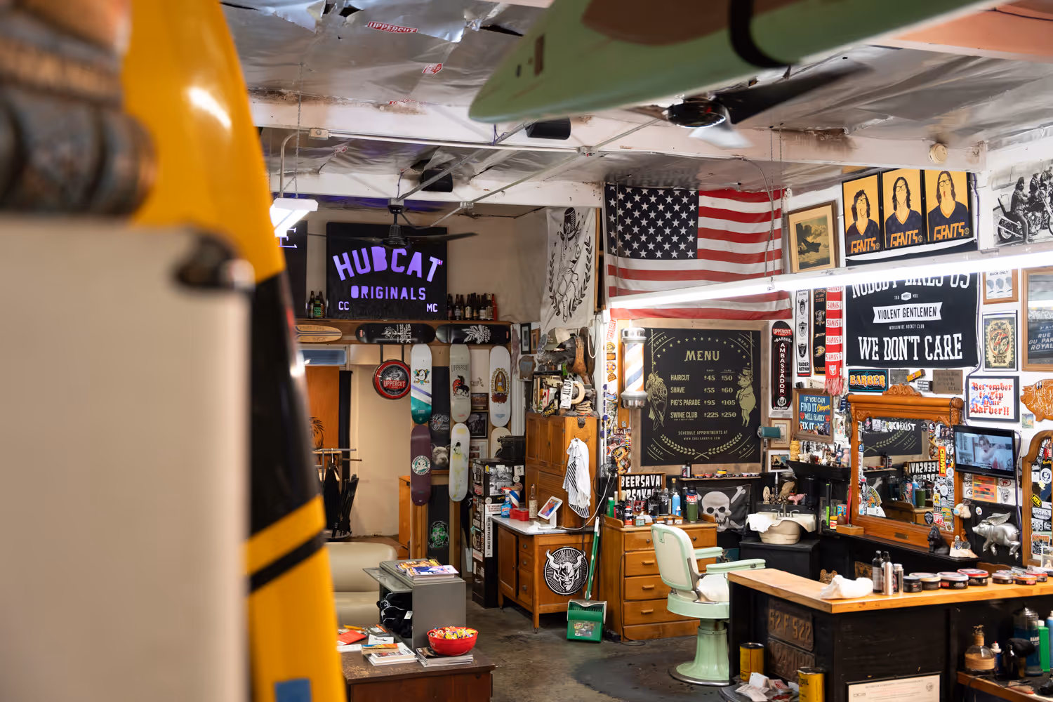 Interior of a vintage barbershop with a green barber chair, wooden counters, an American flag, skateboards on display, and various posters and decor on the walls.