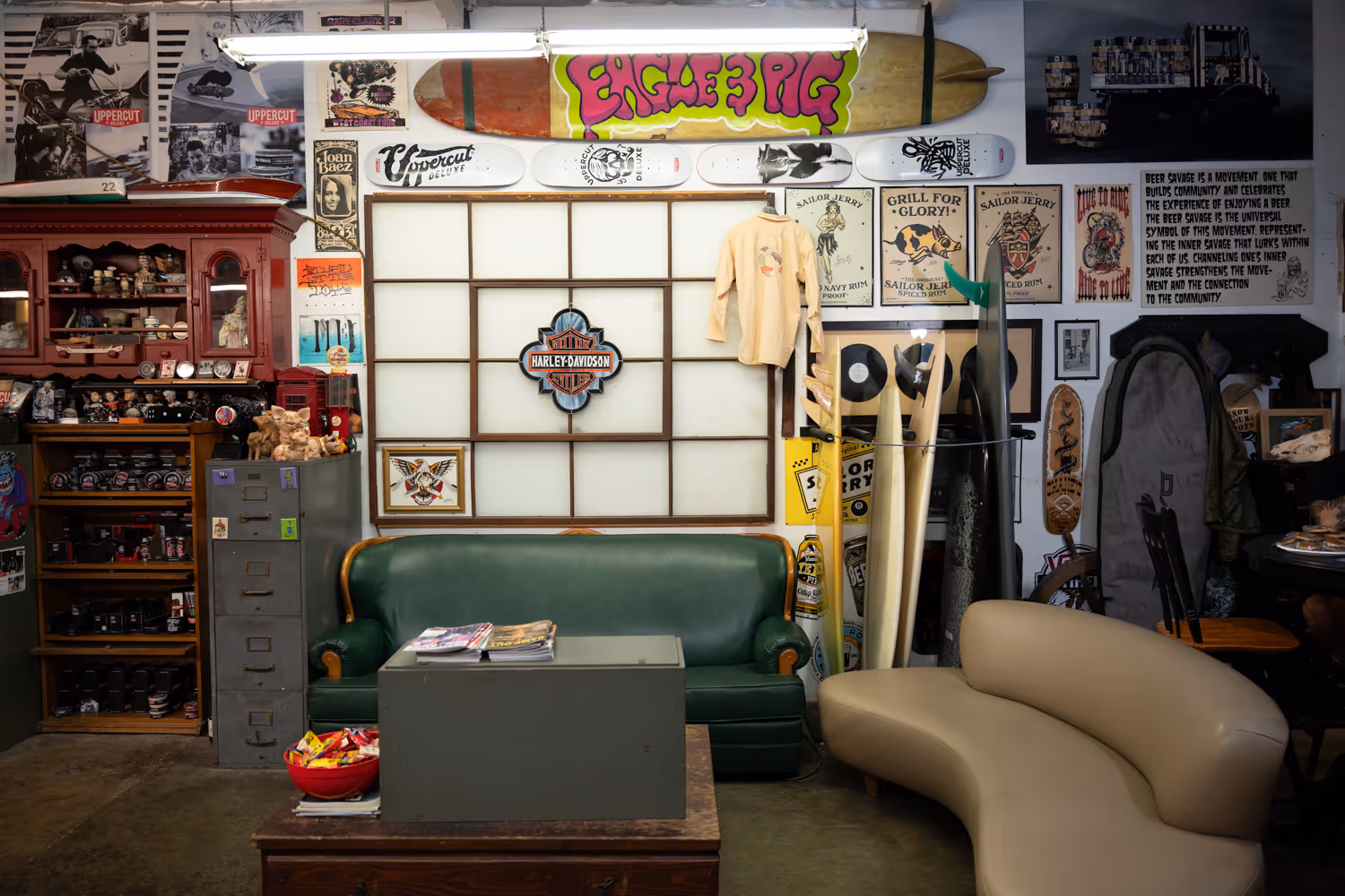 Vintage-style room with green and beige sofas, surfboards, Harley-Davidson sign, and eclectic wall decorations including posters and skateboards.