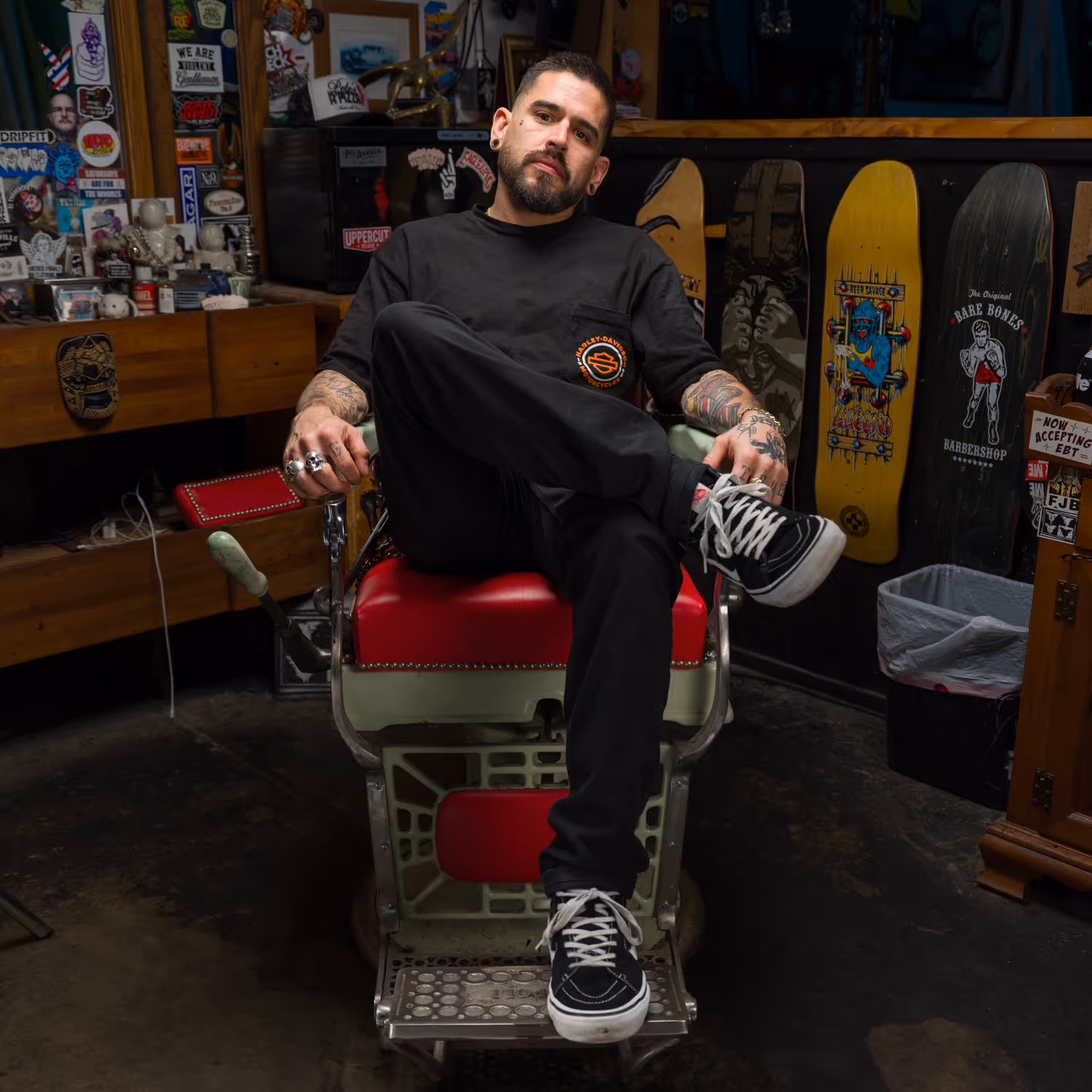 Man with tattoos and rings sitting on a vintage barber chair in a shop decorated with skateboard decks and stickers.