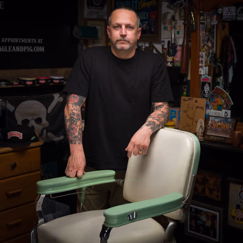 Man with tattooed arms wearing a black t-shirt leaning on a vintage barber chair with pale green armrests inside a tattoo-themed shop.