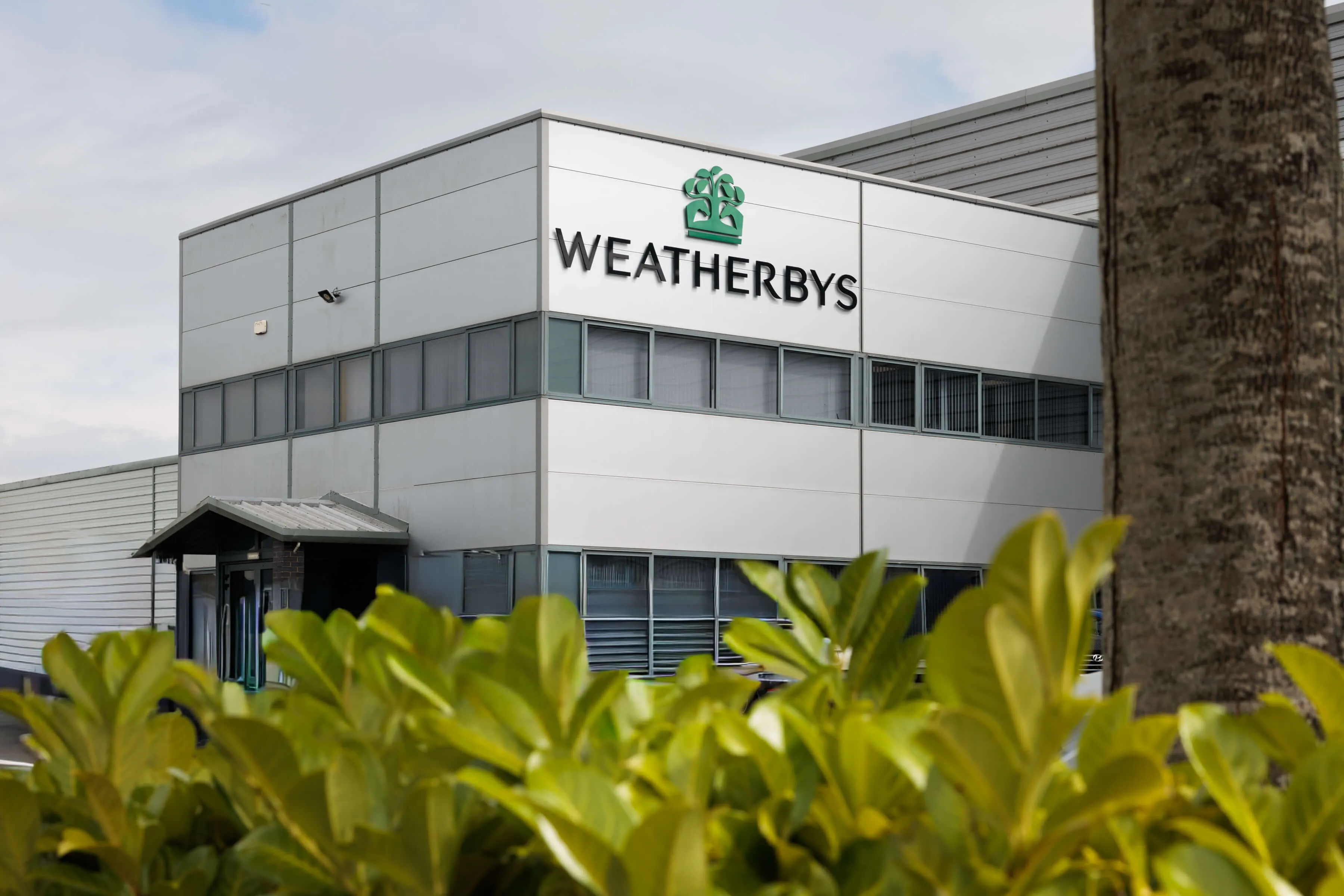 Modern gray commercial building with large windows and Weatherbys logo on the upper facade, partially obscured by green foliage.