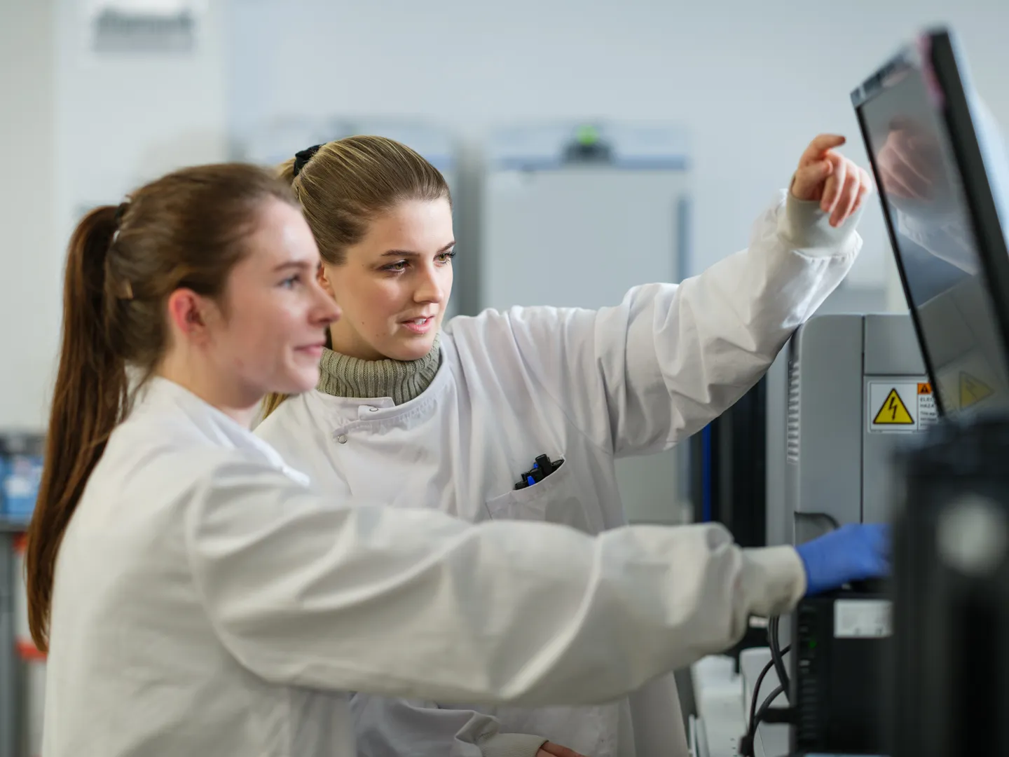Two female scientists in white lab coats operating and discussing data on a laboratory machine screen.