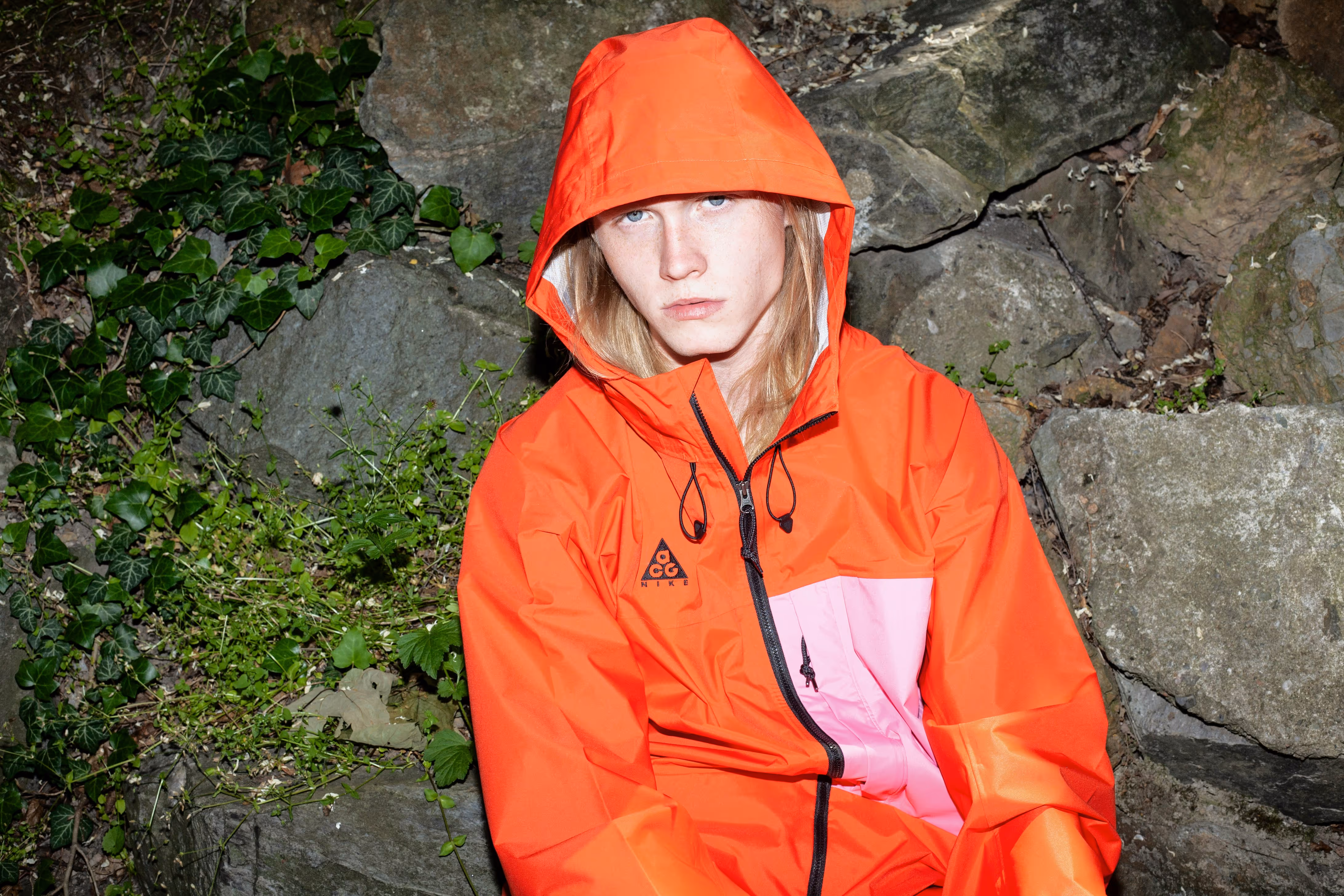 Person with long blond hair wearing an orange and pink Nike ACG hooded jacket sitting against a stone and leafy background.