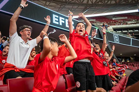 Group of enthusiastic sports fans in red shirts cheering with raised arms in a stadium.