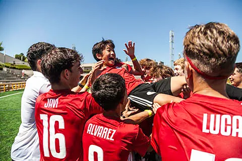 Group of young soccer players celebrating by lifting a teammate in red jersey outdoors under clear blue sky.