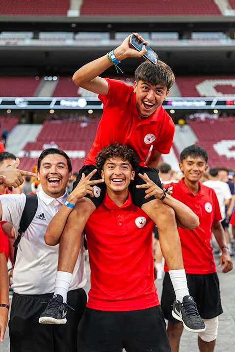 Four young men smiling and posing in a stadium, with one sitting on another’s shoulders while holding a phone.