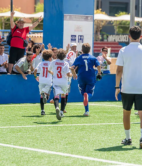 Three children in soccer uniforms running on a field towards cheering spectators.