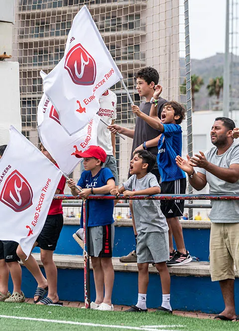 Group of children and a man cheering and waving Euro Soccer Trips flags at a soccer field.