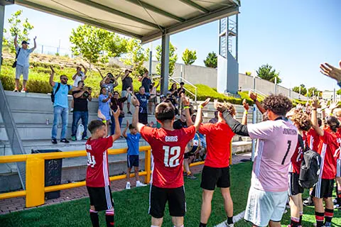 A youth soccer team in red and black uniforms raising their arms toward spectators cheering from bleachers under a covered area.